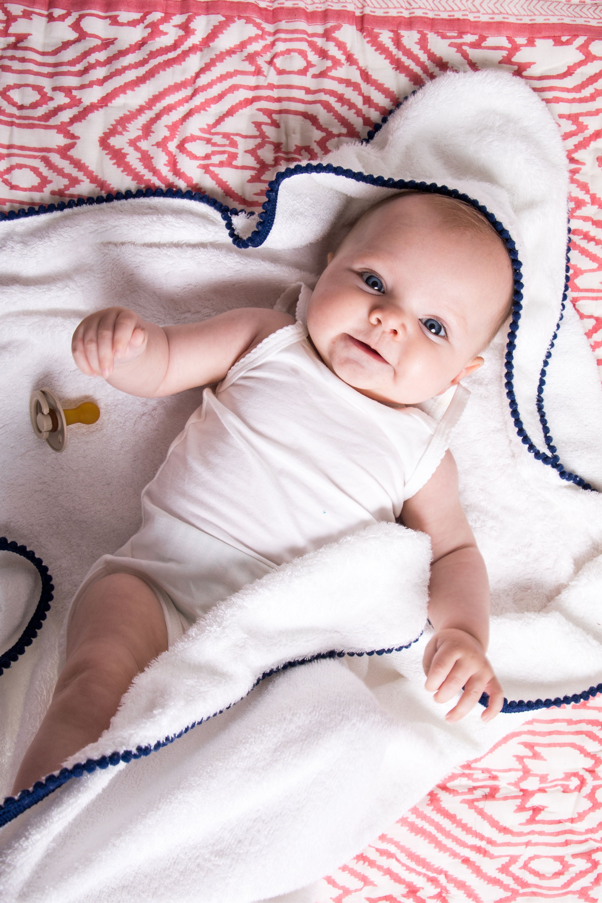 malabar baby white bamboo cotton towel shown on a cute and happy baby. The towel is white and silky soft and has a hood. The towel also has a detailed pom pom trim. The towel is extra large and fits newborns - toddlers 5 years old. Pom Pom colors are show