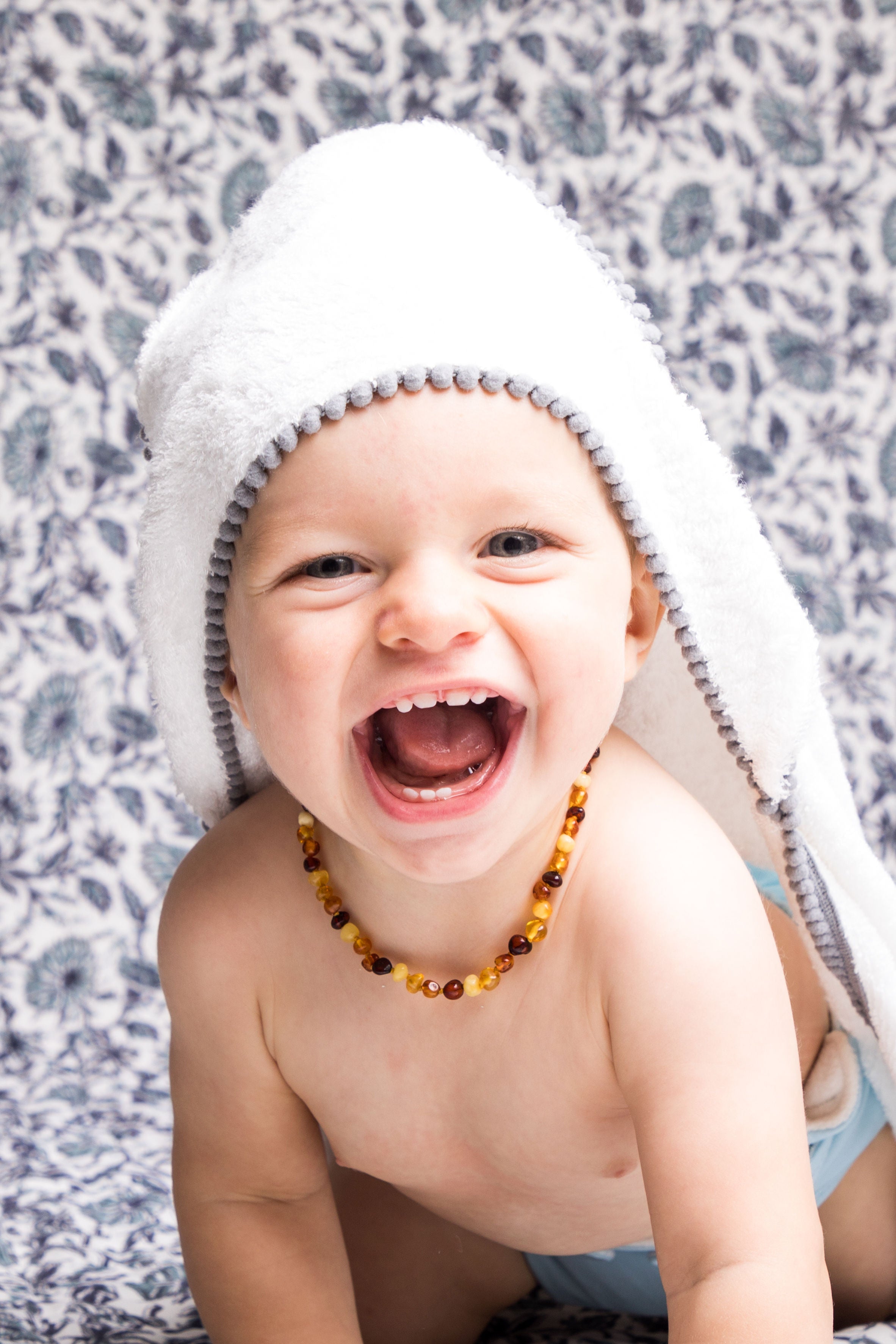 malabar baby white bamboo cotton towel shown on a cute and happy baby. The towel is white and silky soft and has a hood. The towel also has a detailed pom pom trim. The towel is extra large and fits newborns - toddlers 5 years old. Pom Pom colors are show