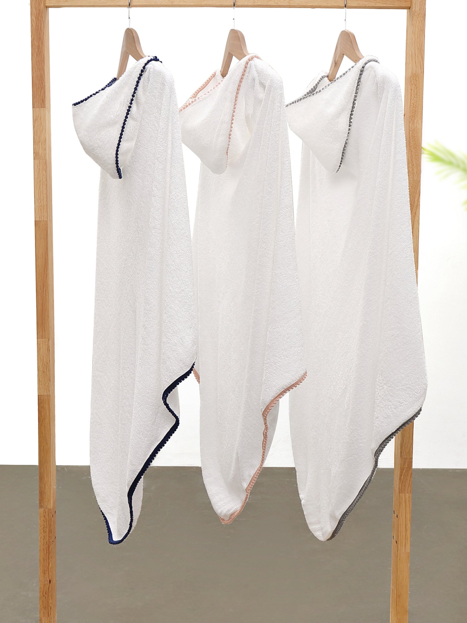 3 white malabar baby bamboo cotton towels hanging on a wooden rack. The towels are white and silky soft and have a hood. They also have a detailed pom pom trim. The towels are extra large and fit from newborns - toddlers 5 years old. Pom Pom colors are; n